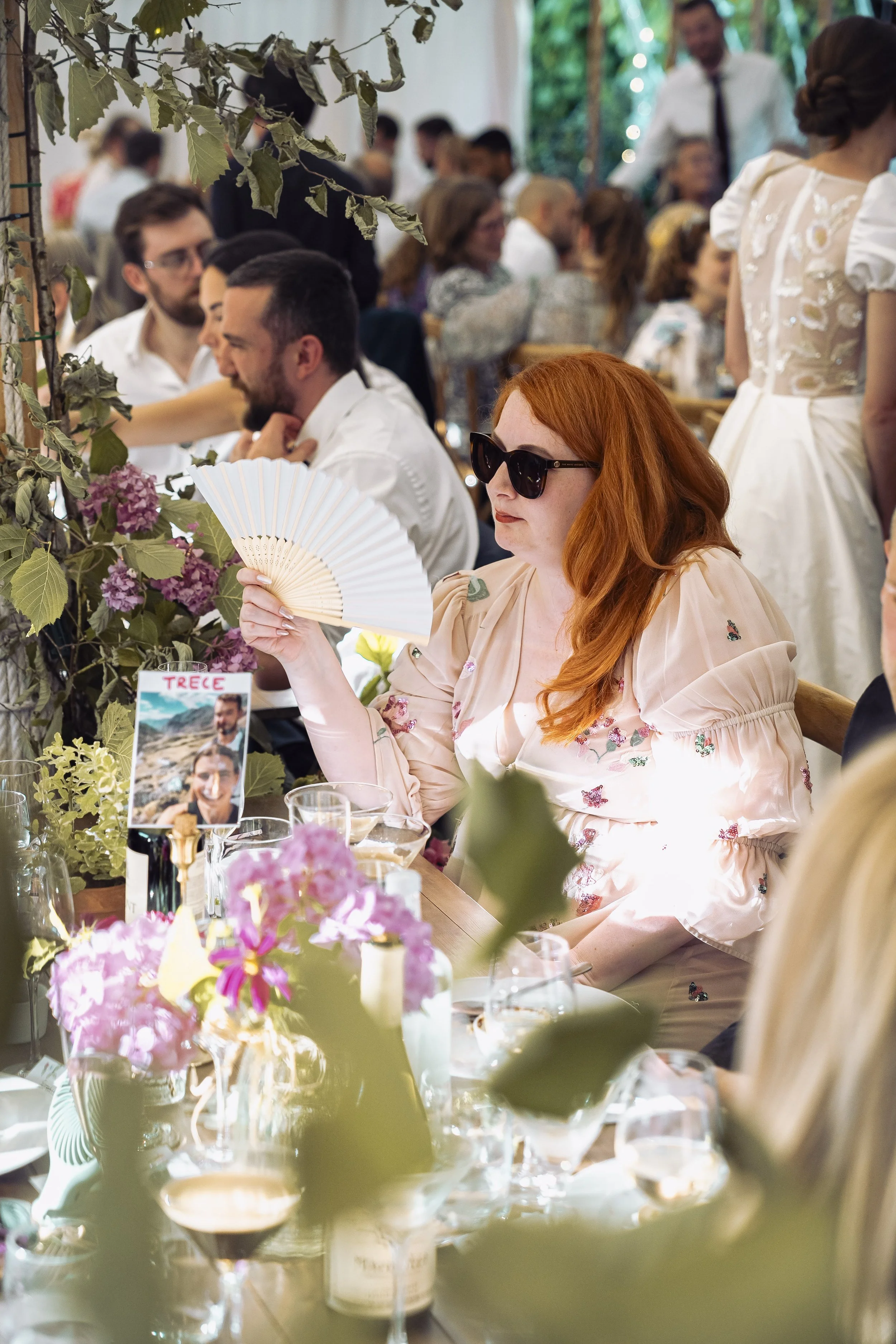 People dining at a festive event with floral centerpieces, greenery, and folded napkins on the table, some guests wearing sunglasses, and a woman with red hair holding a fan.