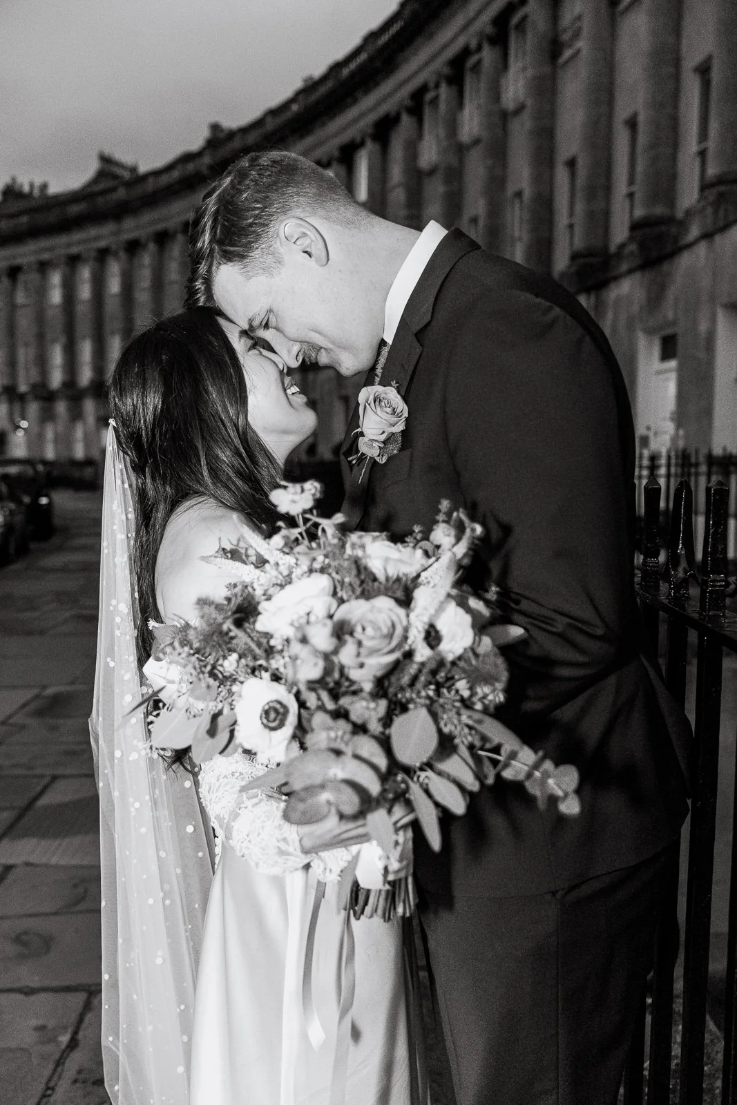 A black and white photo of a bride and groom smiling and touching foreheads, outdoors, with a row of buildings in the background. The bride is holding a bouquet of flowers.
