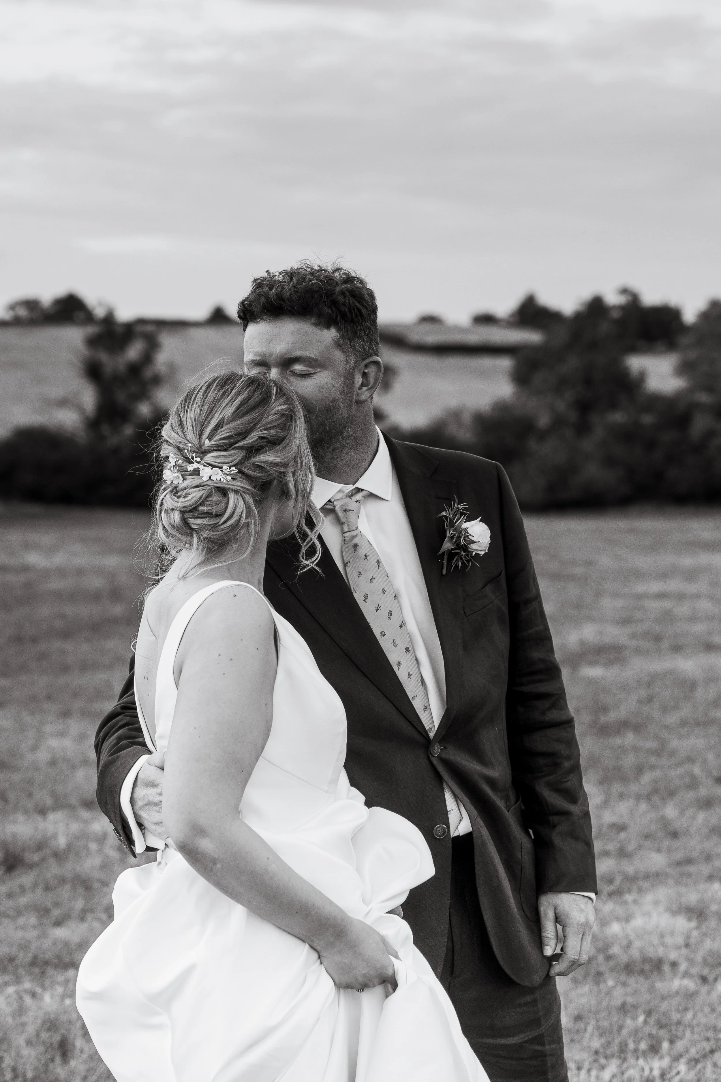 A bride and groom holding each other outdoors in a field, with trees and a cloudy sky in the background, captured in black and white.