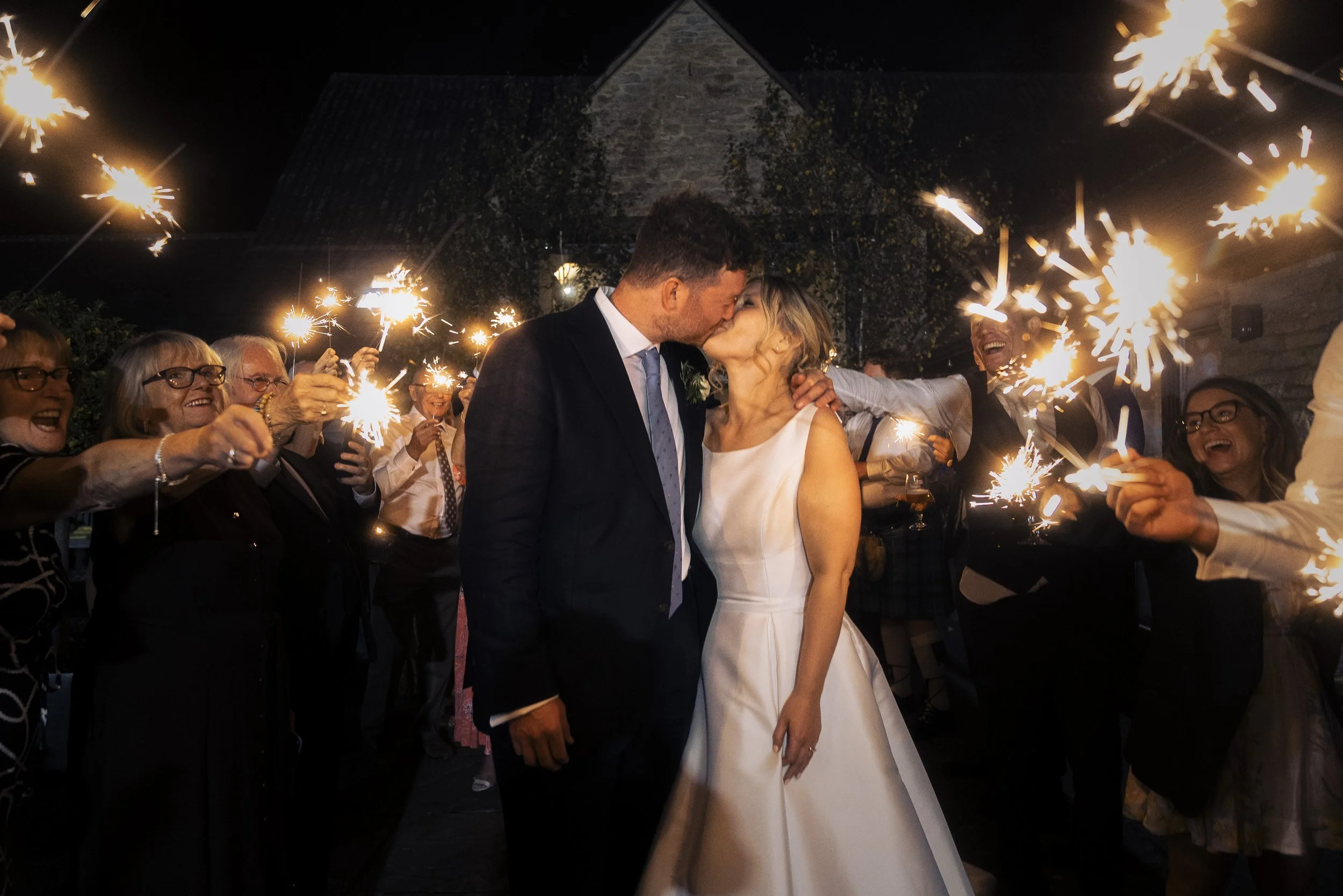 A bride and groom share a kiss at night surrounded by guests holding sparklers during a celebration.