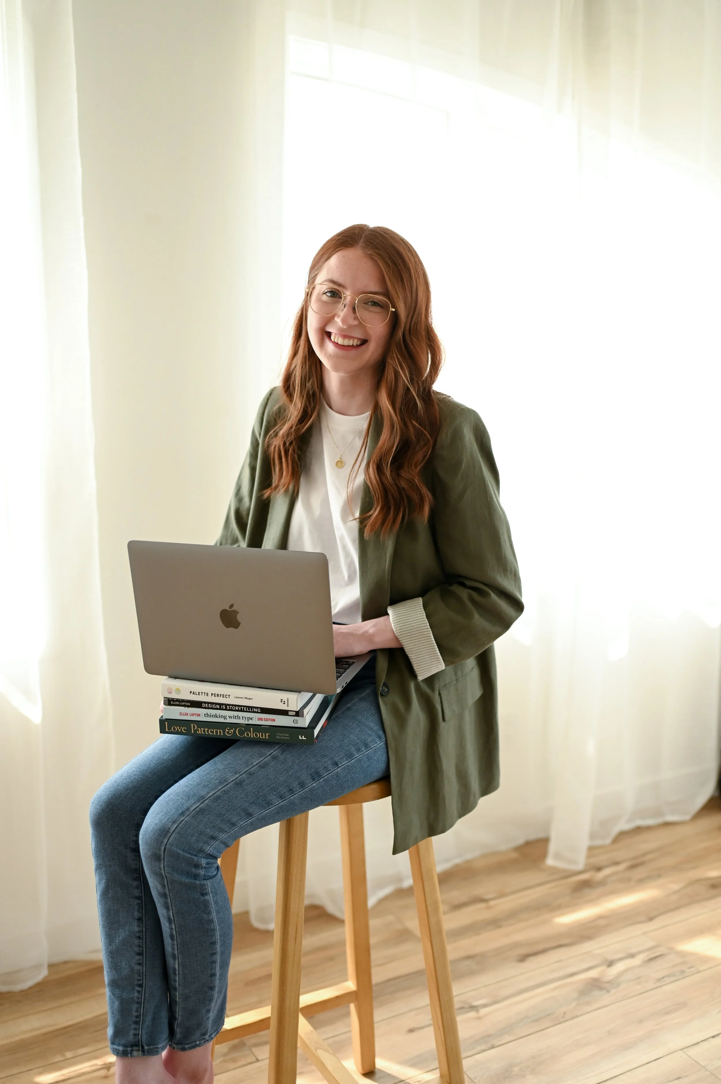 A young woman with long red hair and glasses sitting on a wooden stool, smiling while holding a laptop on her lap and a stack of books.