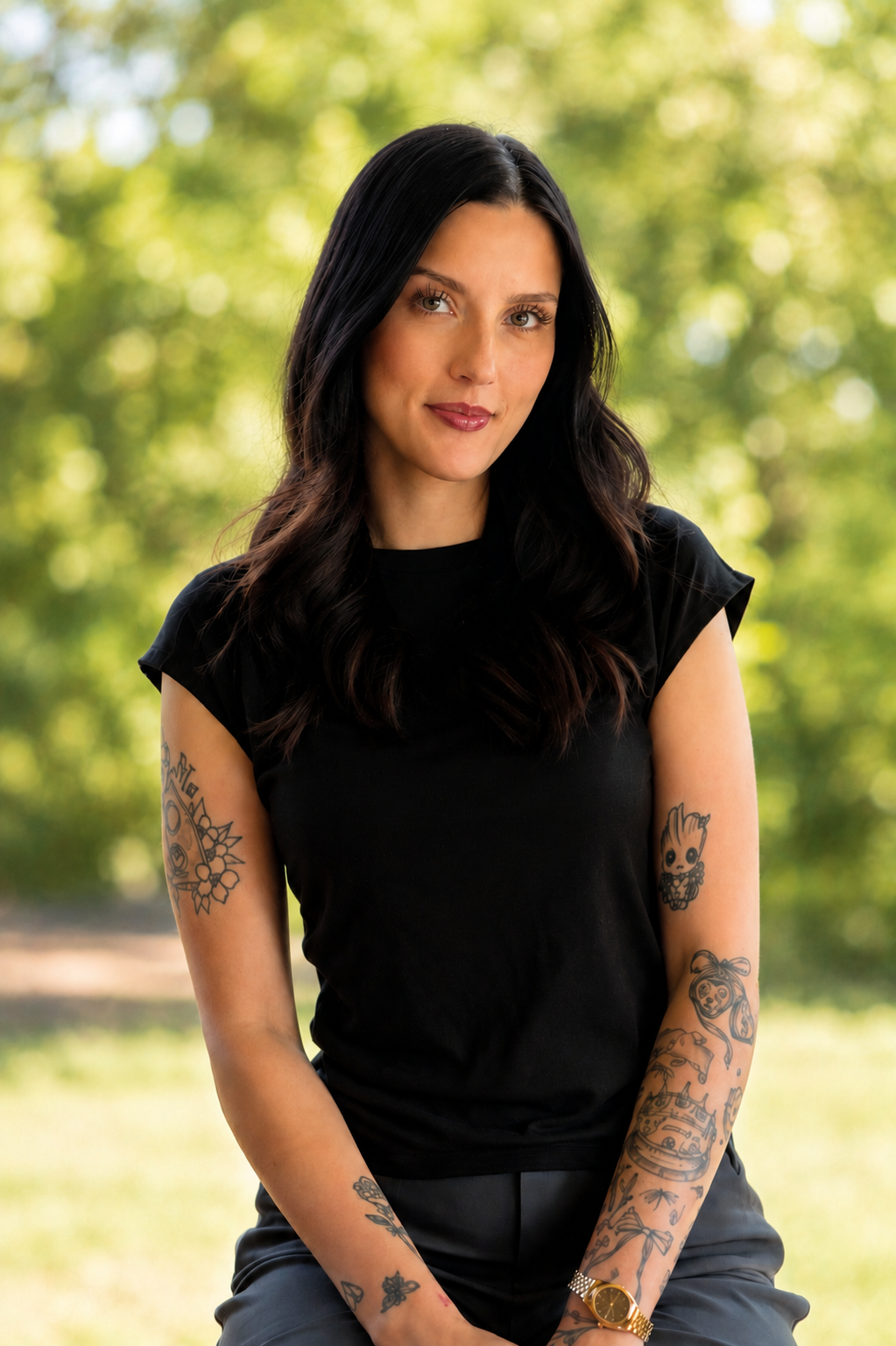 A woman with dark hair and tattoos on her arms, wearing a black shirt, sitting outdoors with a blurred green nature background.