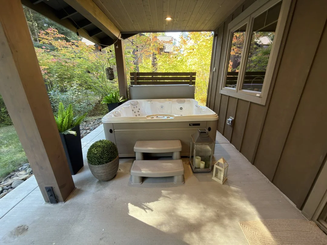 Outdoor covered porch with hot tub, potted plants, lanterns, and candles, surrounded by trees with fall foliage.
