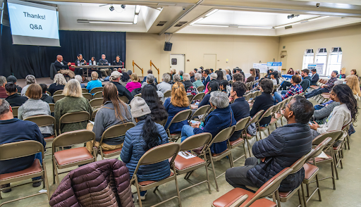 Image of a crowd of people facing a stage. On stage are several people sitting at a table underneath a sign that says 'Thanks! Q&A'