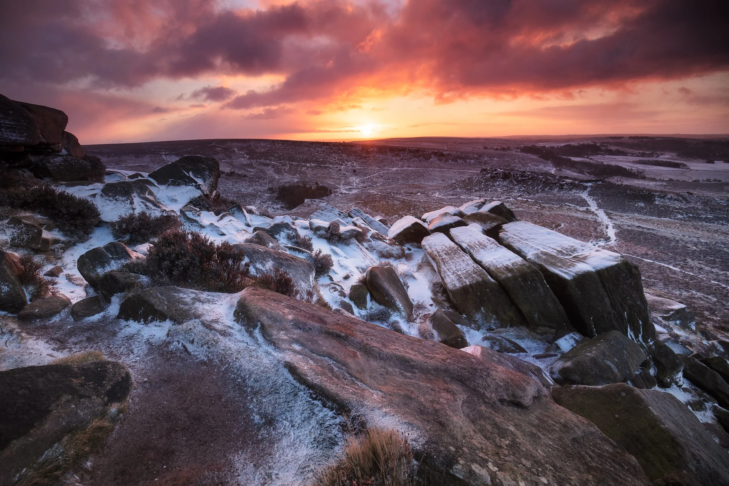 A scenic winter landscape during sunset with snow-covered rocks in the foreground and rolling hills in the distance under a colorful sky with clouds.