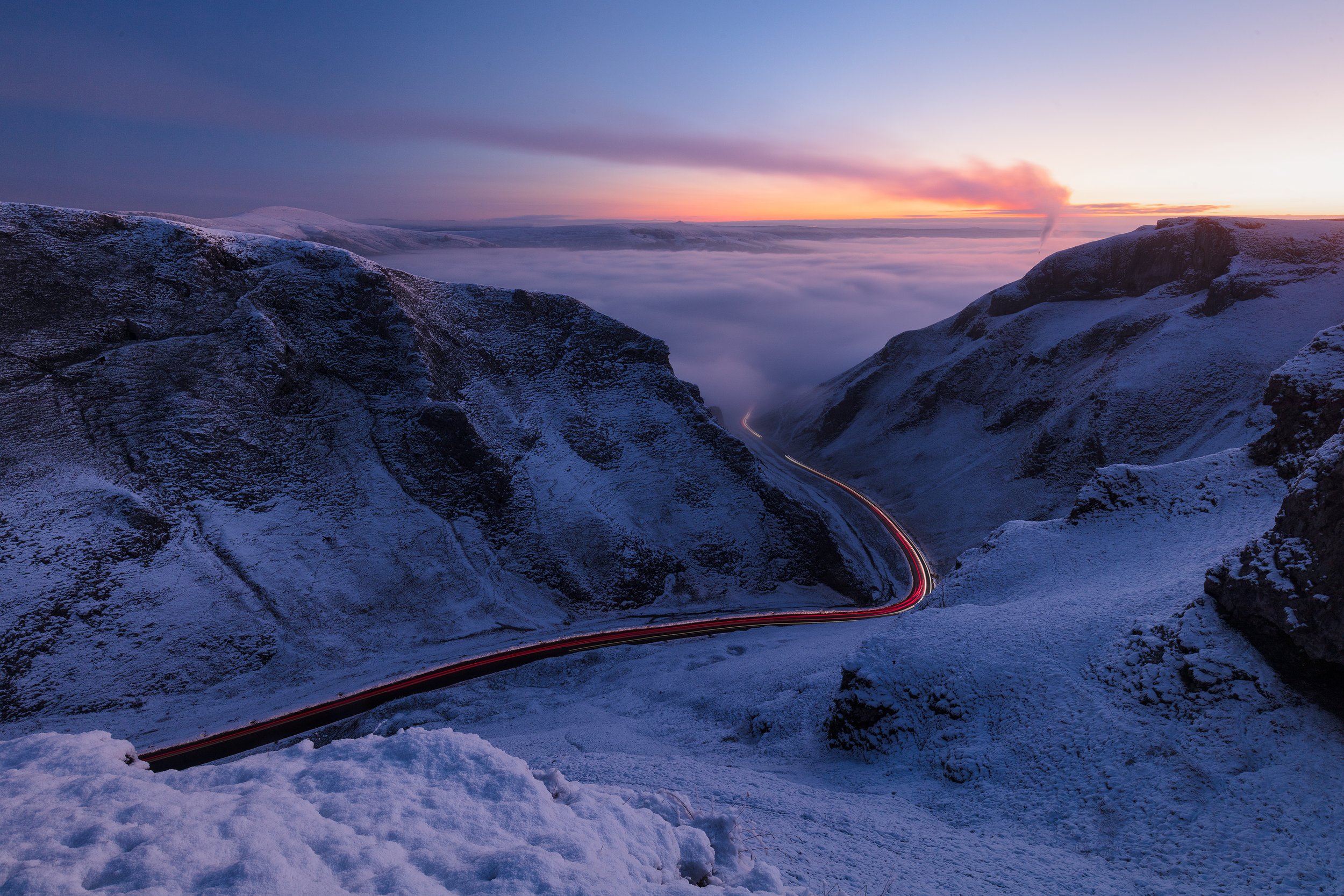 Snow-covered mountain landscape at sunset with winding road and streaks of vehicle lights, overcast sky with horizon glow.