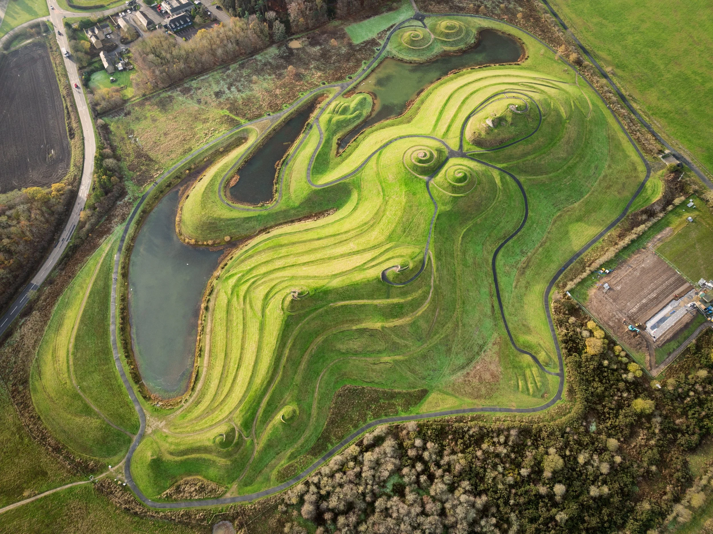Aerial view of a large, artistically landscaped park with winding pathways, a pond, and lush green grass with intricate swirled patterns.