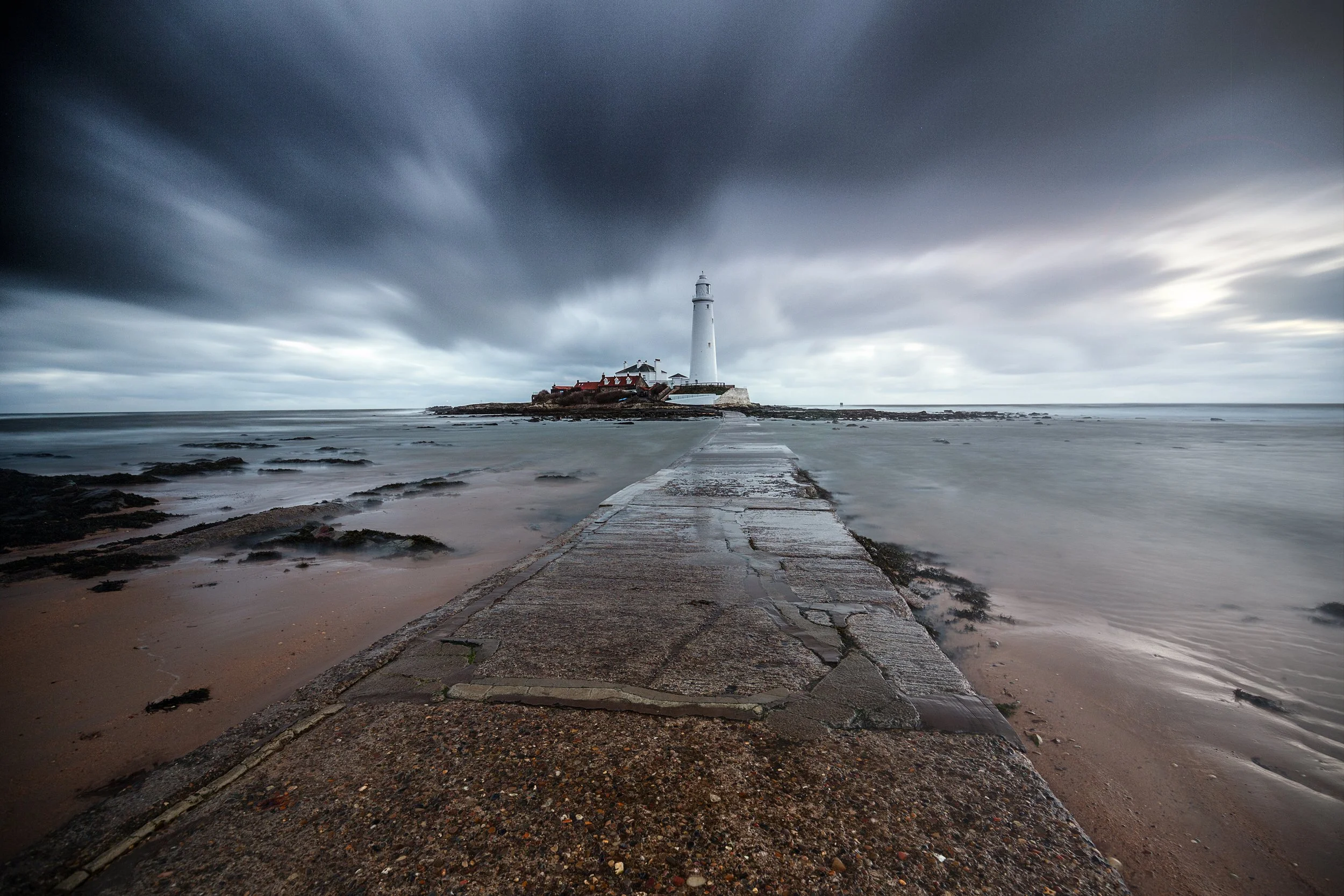 A lighthouse on a small island connected by a concrete pathway, with dark clouds overhead and a cloudy sky.