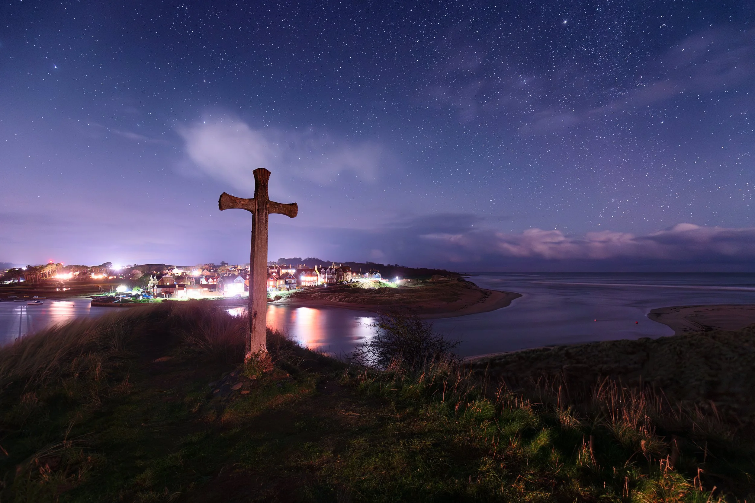Nighttime view of a coastal town with a large wooden cross on a hill in the foreground, starry sky, and illuminated buildings reflecting on the water.