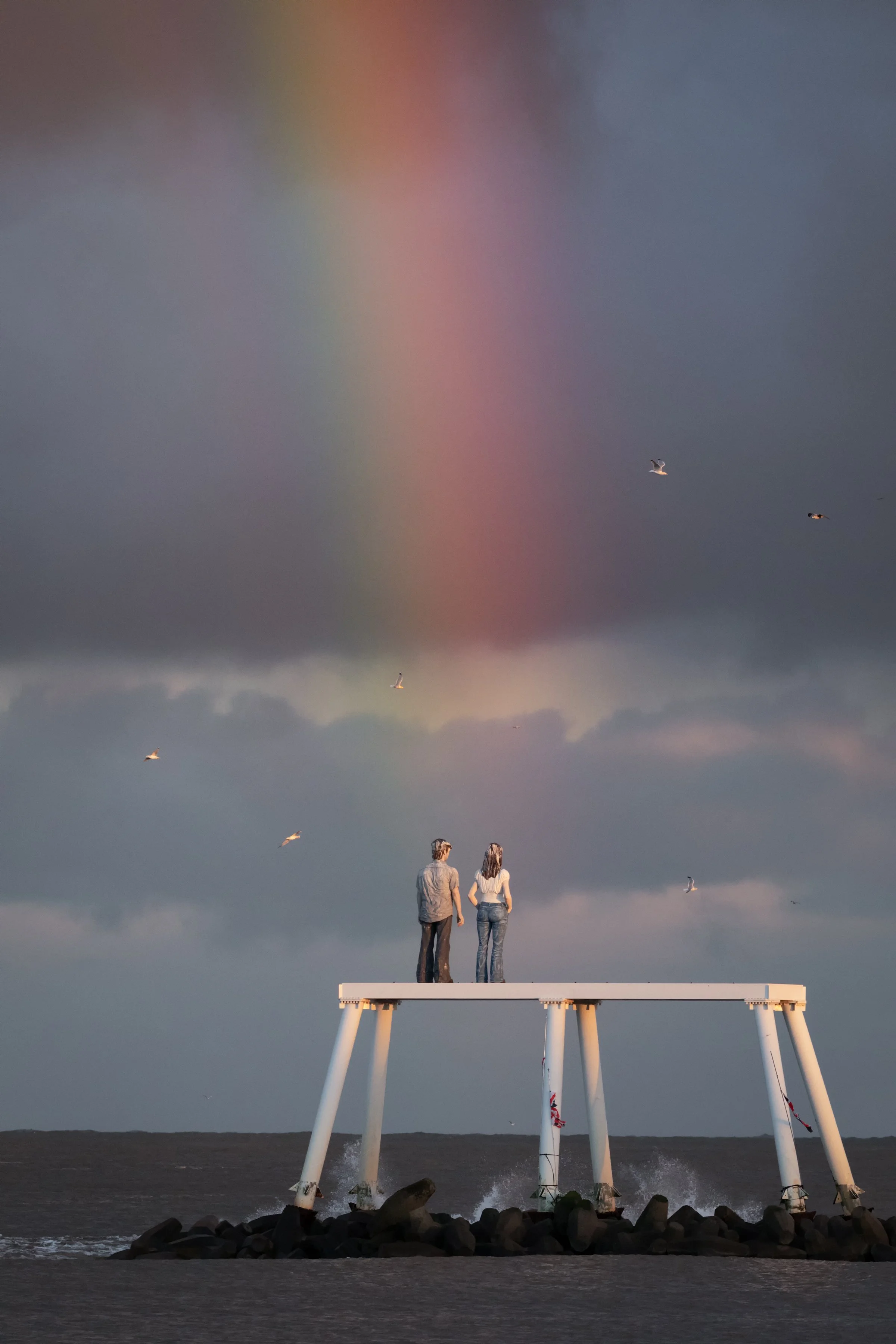 Two people stand on a small platform at the beach, with a rainbow and dark storm clouds in the background and seagulls flying overhead.
