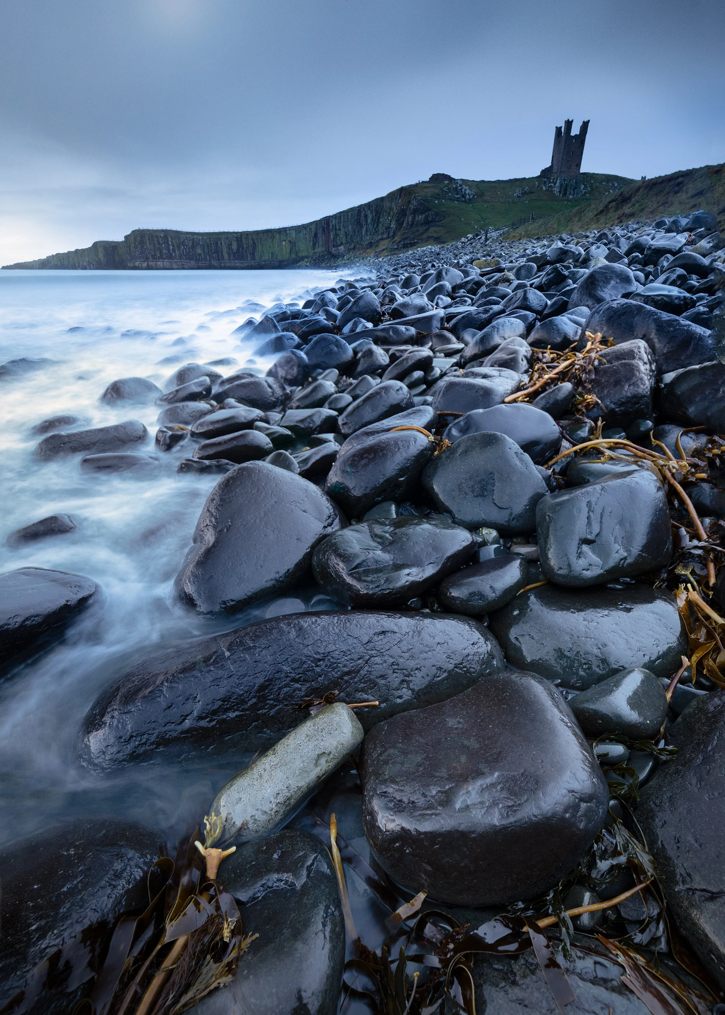 A rocky beach with wet, smooth stones and seaweed, with a castle on a hill in the background and cloudy sky.