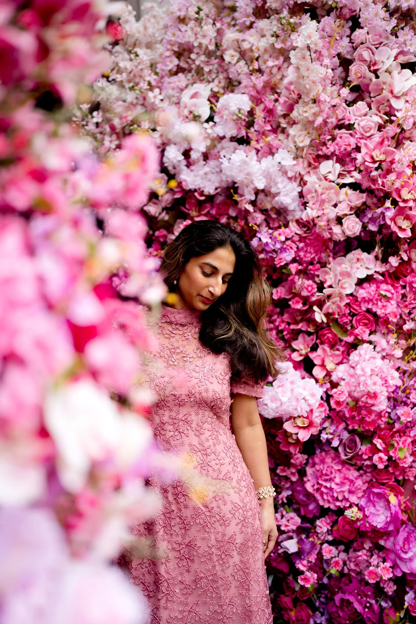 Woman in pink lace dress standing amidst a wall of vibrant pink and white flowers.