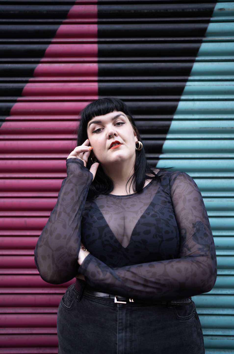 A woman with black hair and bangs posing in front of a colorful striped metal shutter. She is wearing a sheer black patterned top and dark jeans, with a confident expression.