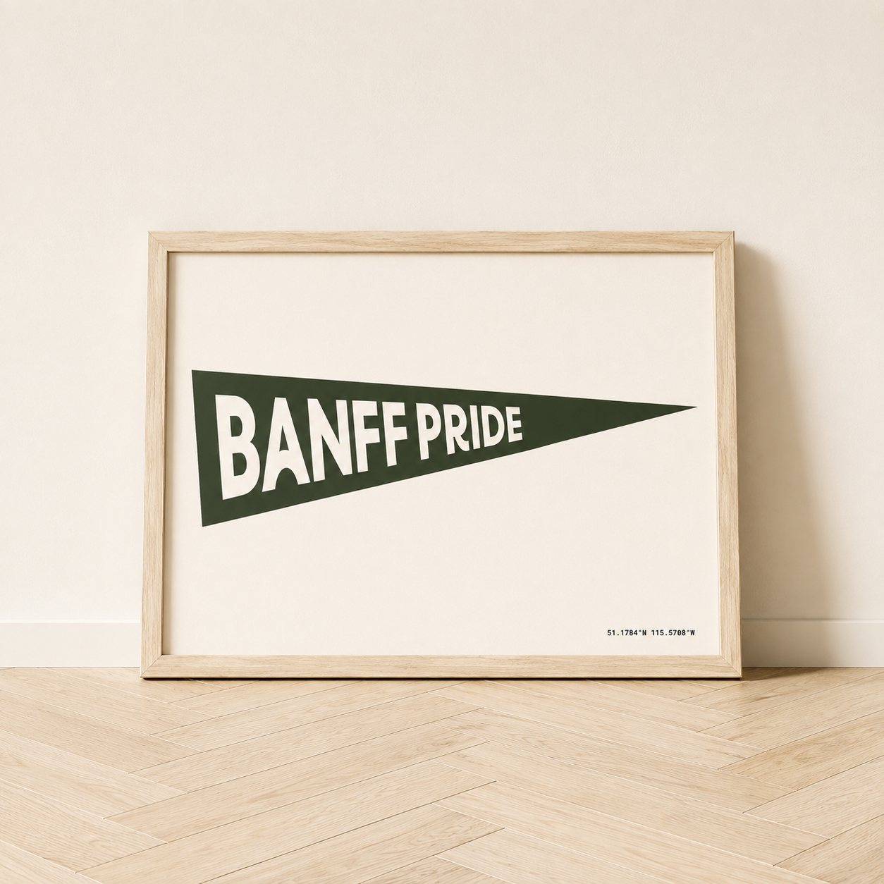 Banff Pride Pennant Flag art print in a natural wood frame, featuring a dark spruce green triangular pennant with white “Banff Pride” lettering and Banff coordinates on a cream background.