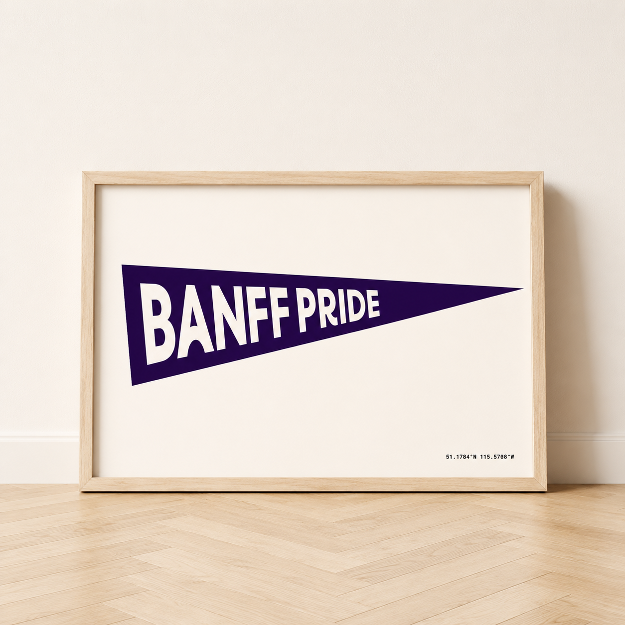 Banff Pride Pennant Flag art print in a natural wood frame, featuring a dark denim blue triangular pennant with white “Banff Pride” lettering and Banff coordinates on a cream background.