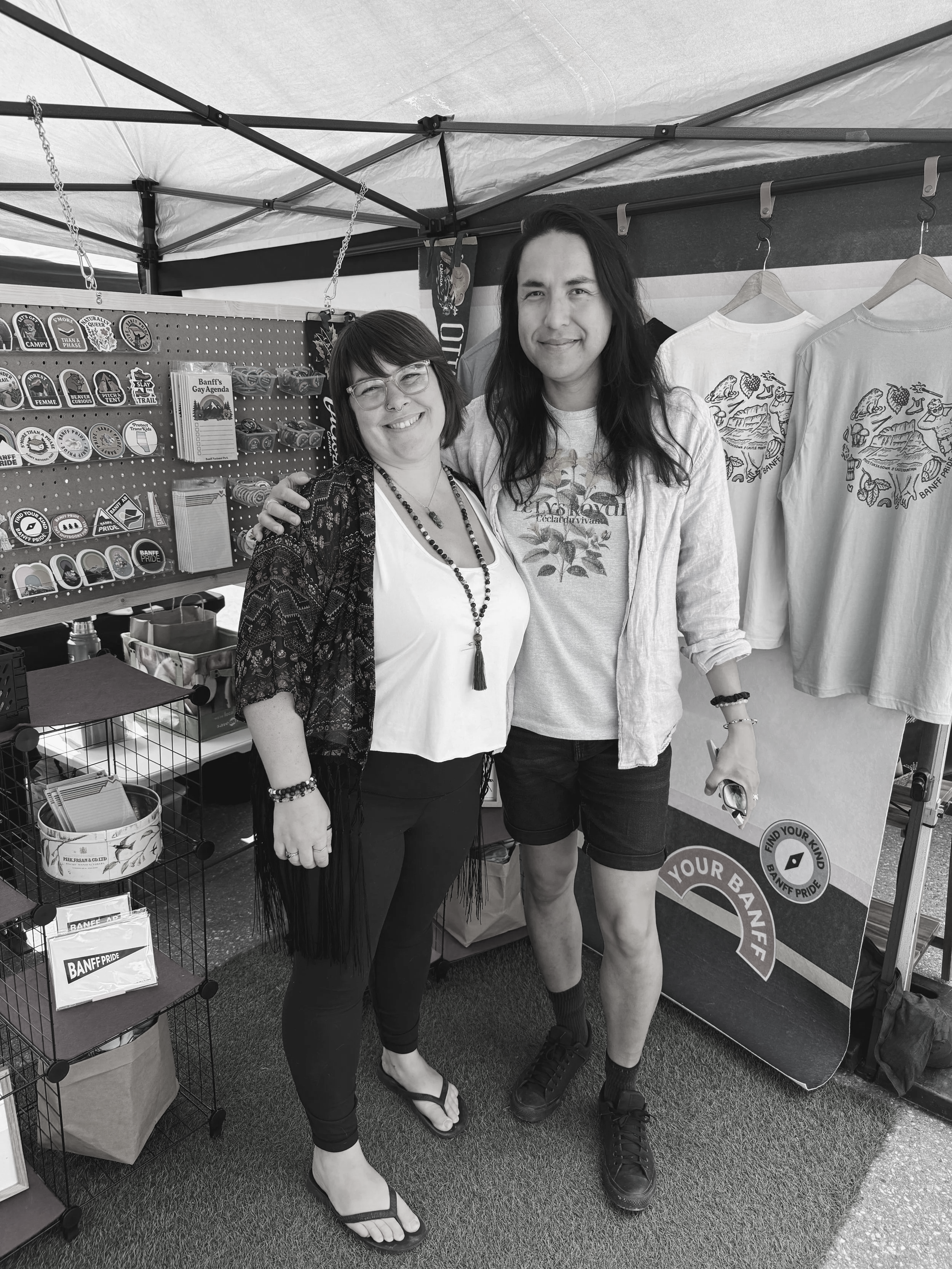 Two people standing inside a tent at a booth with various patches, stickers, and T-shirts on display. The woman on the left has shoulder-length hair, glasses, and is wearing a patterned cardigan over a white top with necklaces. The man on the right has long dark hair, a light-colored shirt over a graphic tee, and shorts. They are smiling and posing for the photo.