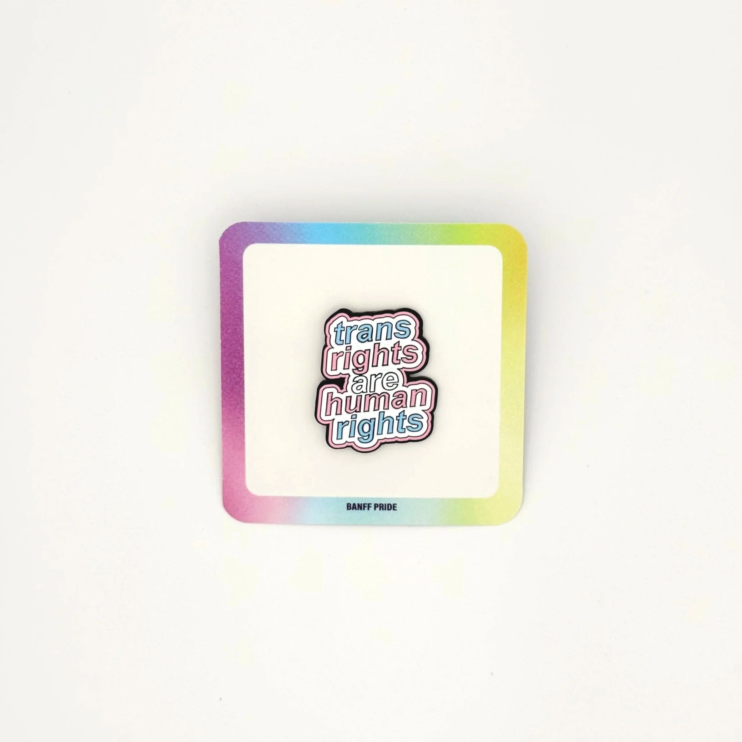 Enamel pin reading “trans rights are human rights” in blue, pink, and white lettering, displayed on a square Banff Pride backing card with a rainbow-gradient border.