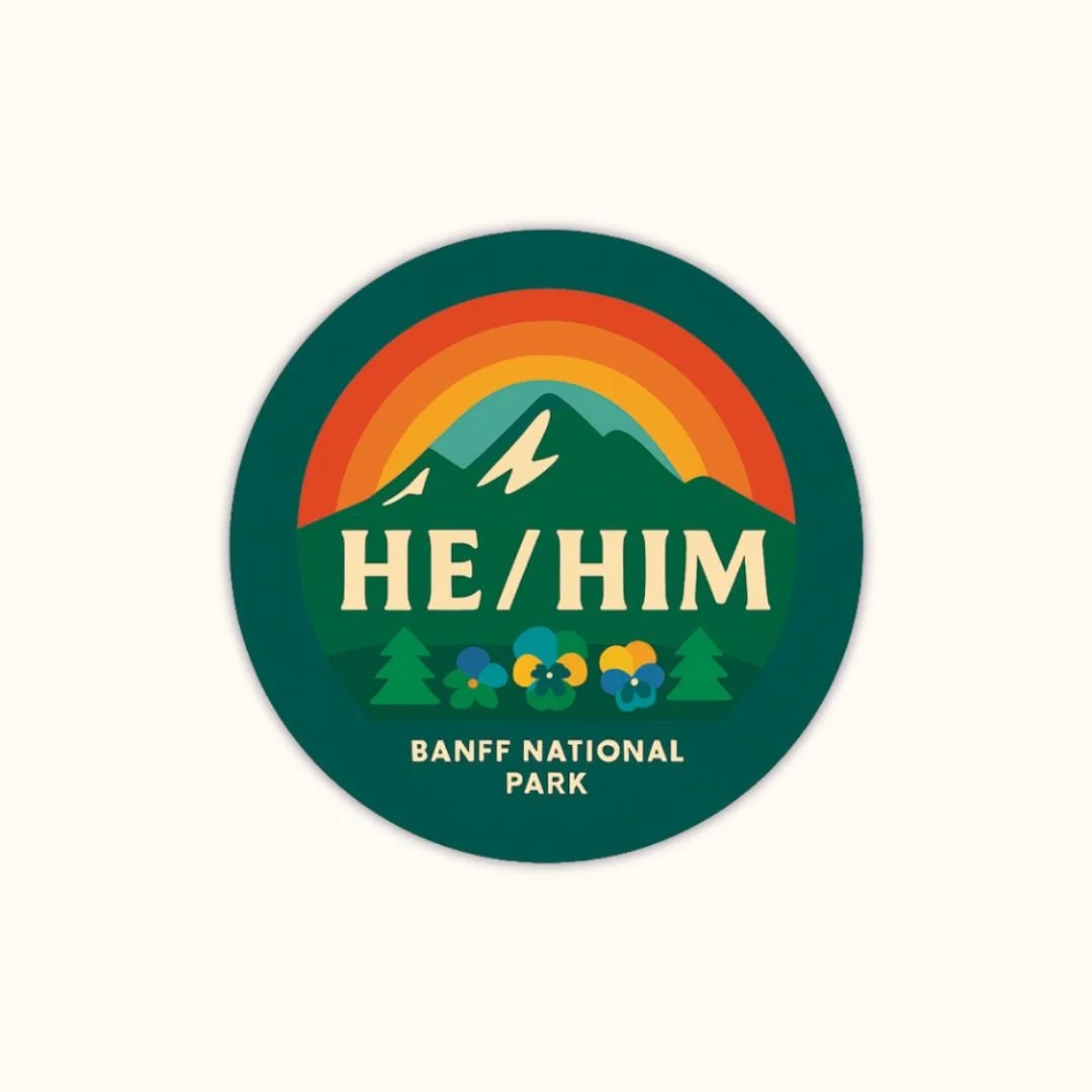Round super glossy “He/Him” button with a rainbow over mountain peaks, small trees and flowers, and the text “Banff National Park.”