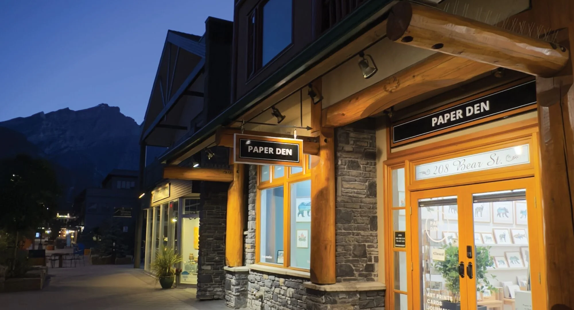 Exterior of a store called Paper Den at 208 Bear Street, with wooden and stone facade, illuminated sign, and large display windows showing artwork inside, during evening with mountain backdrop.