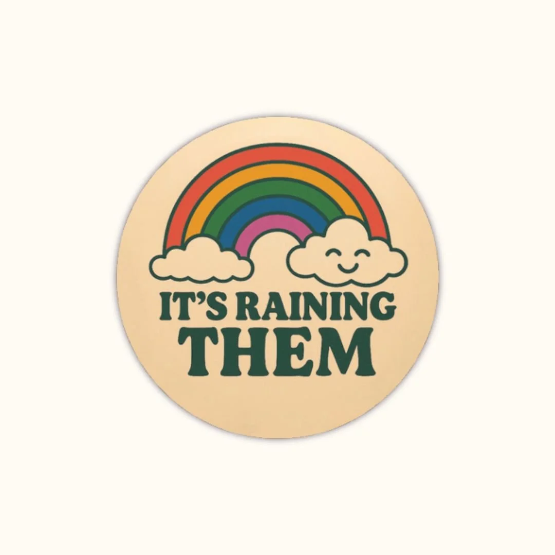 Round super glossy button reading “IT’S RAINING THEM” with a rainbow and two smiling clouds above the text.