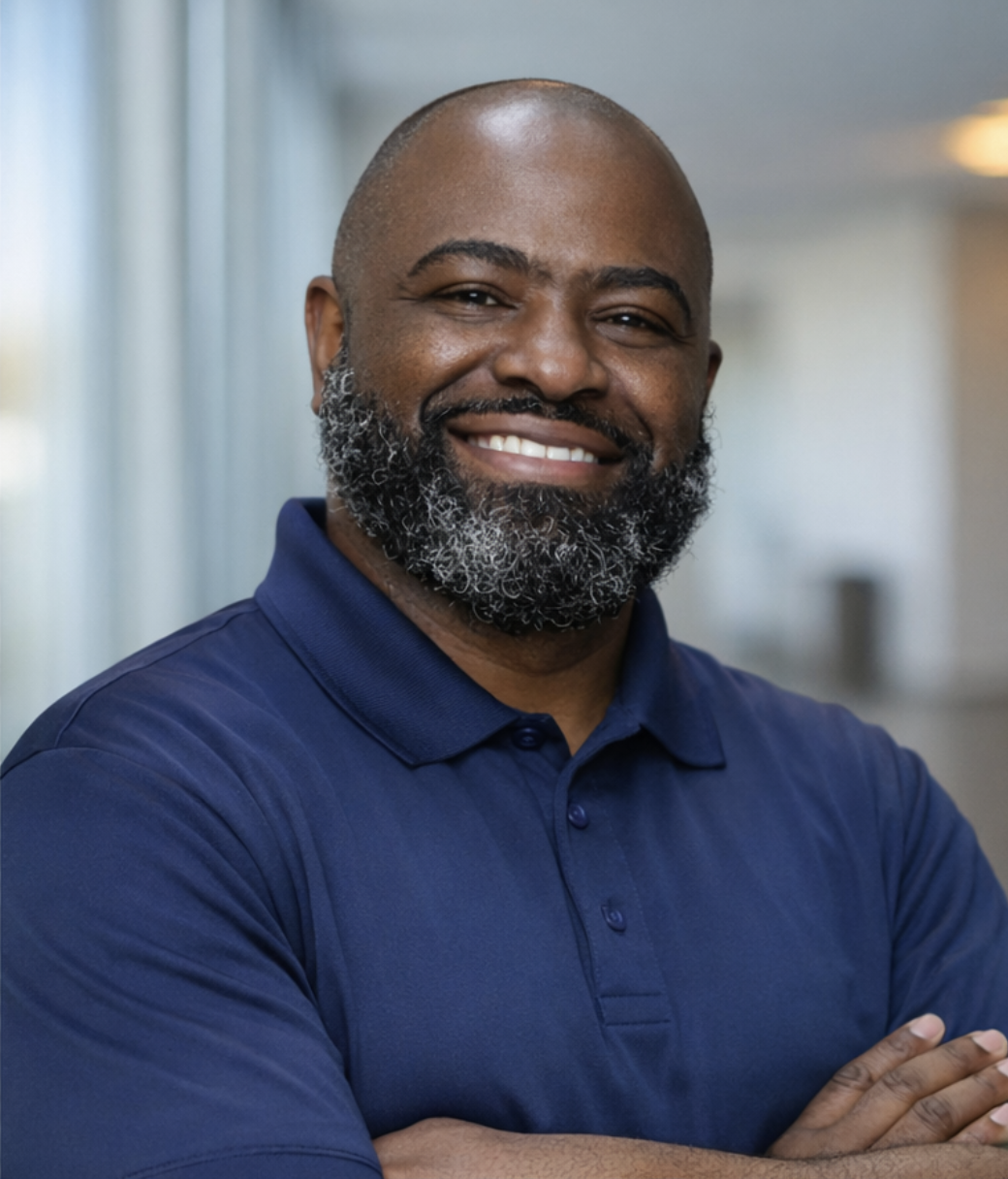 A smiling Black man with a beard wearing a dark blue polo shirt, sitting in a room with warm-colored curtains in the background.