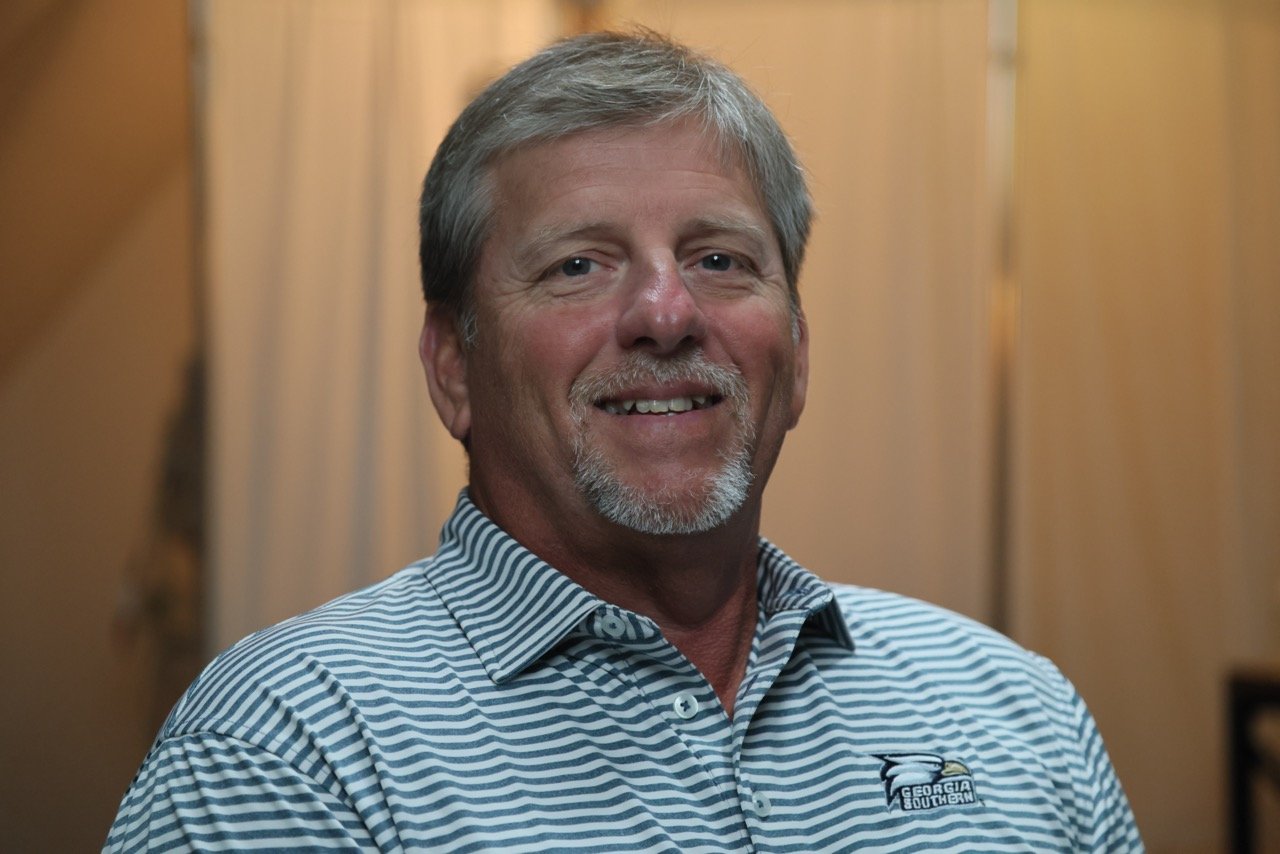 Smiling man with gray hair and beard wearing a Georgia Southern University shirt, standing indoors with wooden panel walls in the background.