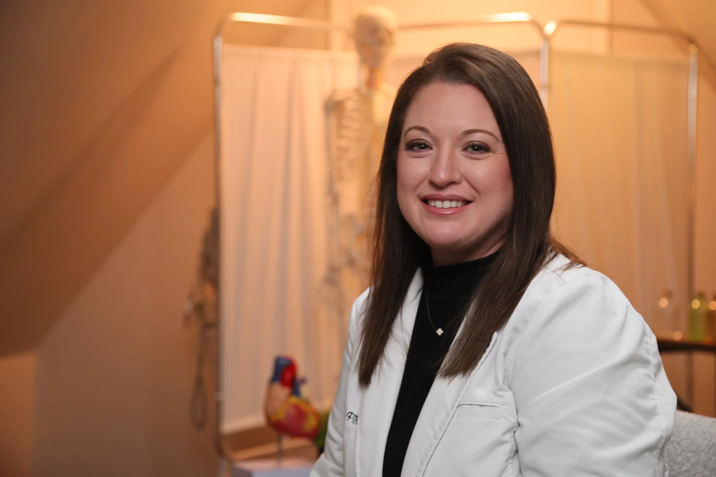 A woman with straight brown hair, wearing a white medical coat and a black top, smiling at the camera in a medical setting with a skeleton model and medical supplies in the background.