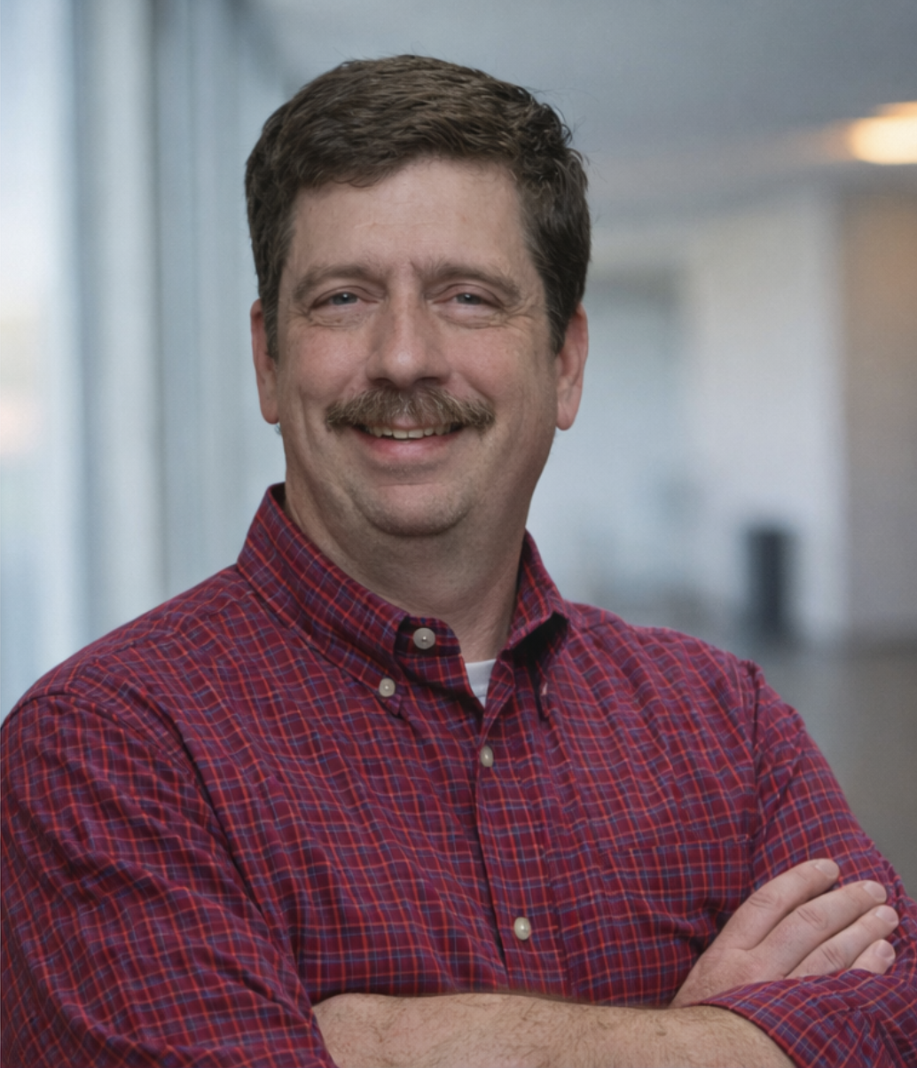 A man with dark hair and a mustache is smiling while sitting on a chair. In the background, there is a lamp with a yellow shade and some decorative items on a shelf, with warm lighting creating a cozy atmosphere.