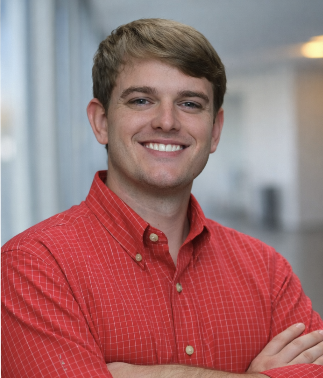 A young man with light brown hair, wearing a red checkered shirt, smiling and looking at the camera.