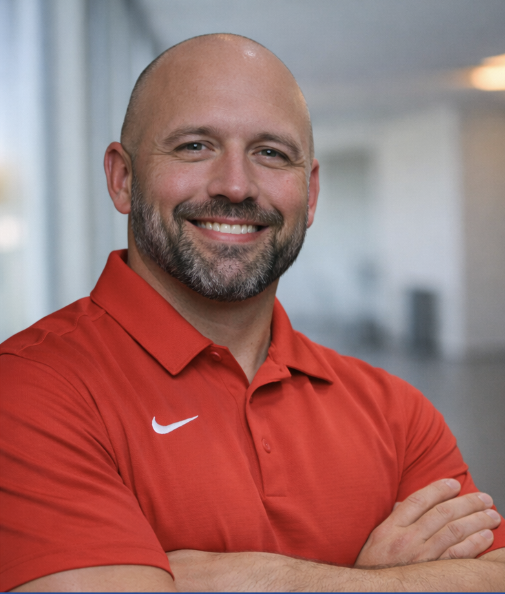 A smiling man with a beard and shaved head wearing a red Nike polo shirt, sitting indoors in a well-lit room with a lamp and decorative items in the background.