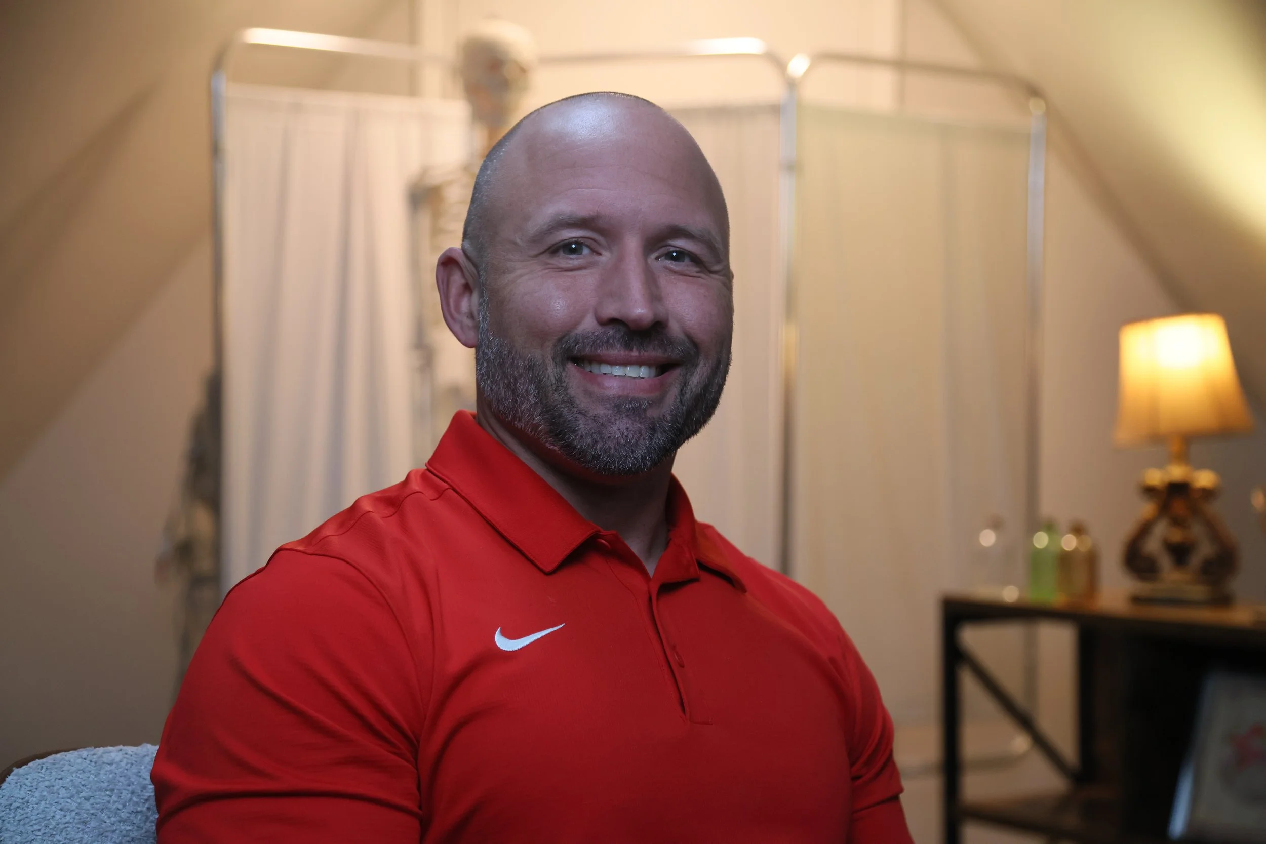 A smiling man with a beard and shaved head wearing a red Nike polo shirt, sitting indoors in a well-lit room with a lamp and decorative items in the background.