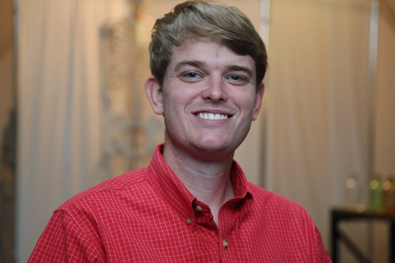 A young man with light brown hair, wearing a red checkered shirt, smiling and looking at the camera.