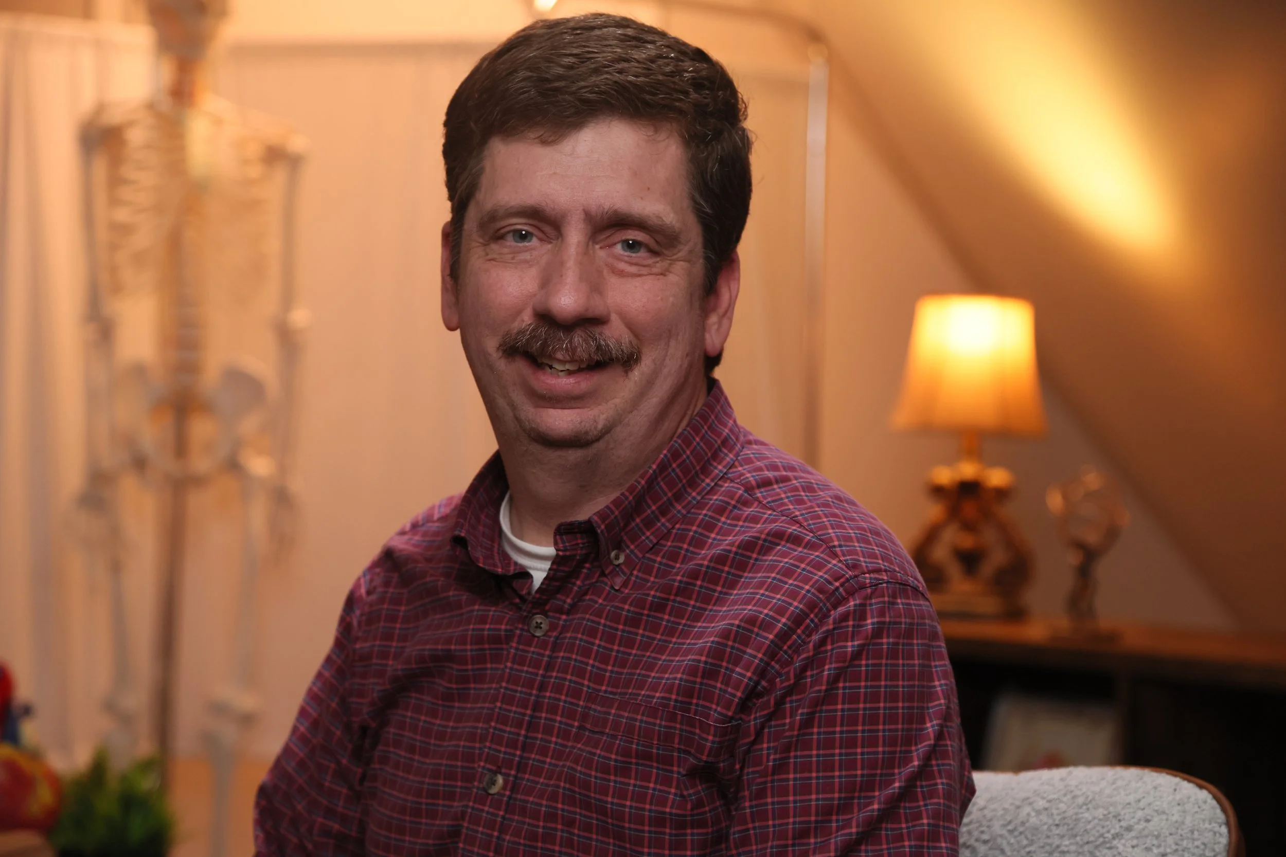A man with dark hair and a mustache is smiling while sitting on a chair. In the background, there is a lamp with a yellow shade and some decorative items on a shelf, with warm lighting creating a cozy atmosphere.