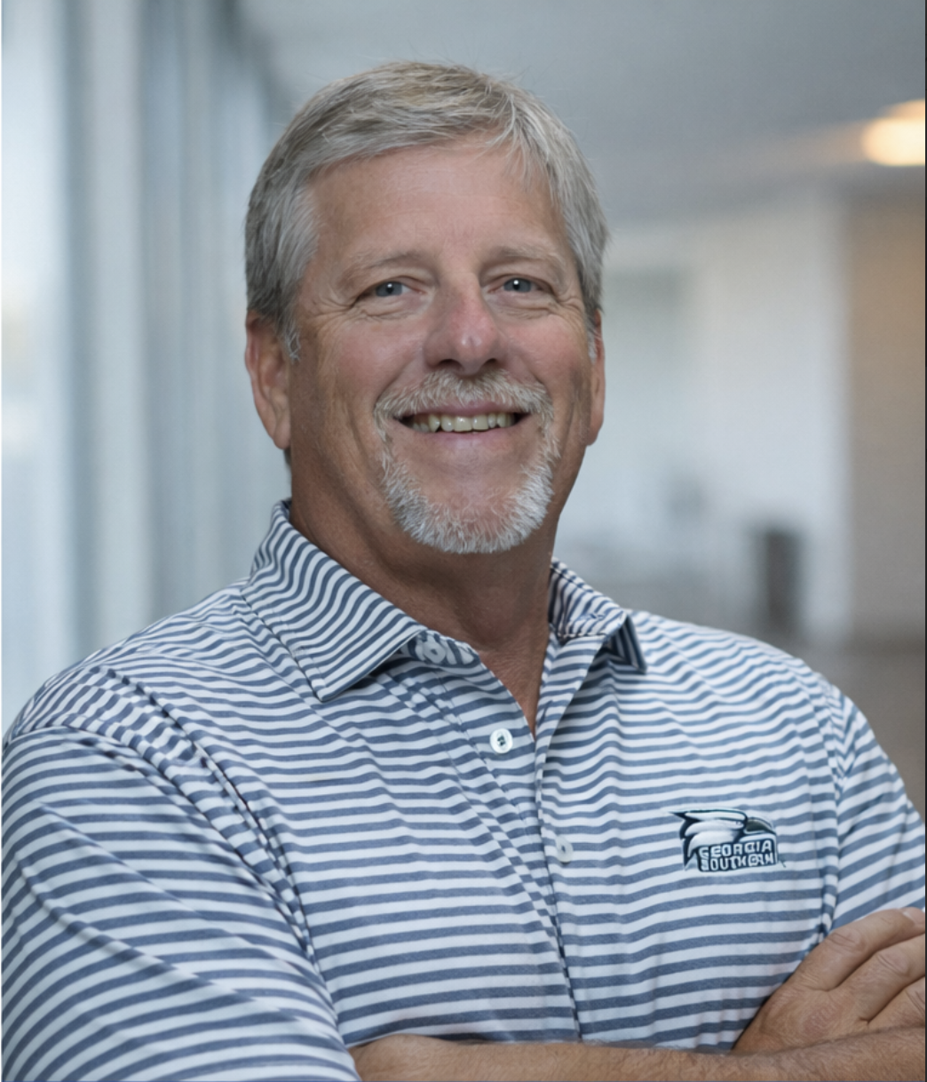 Smiling man with gray hair and beard wearing a Georgia Southern University shirt, standing indoors with wooden panel walls in the background.
