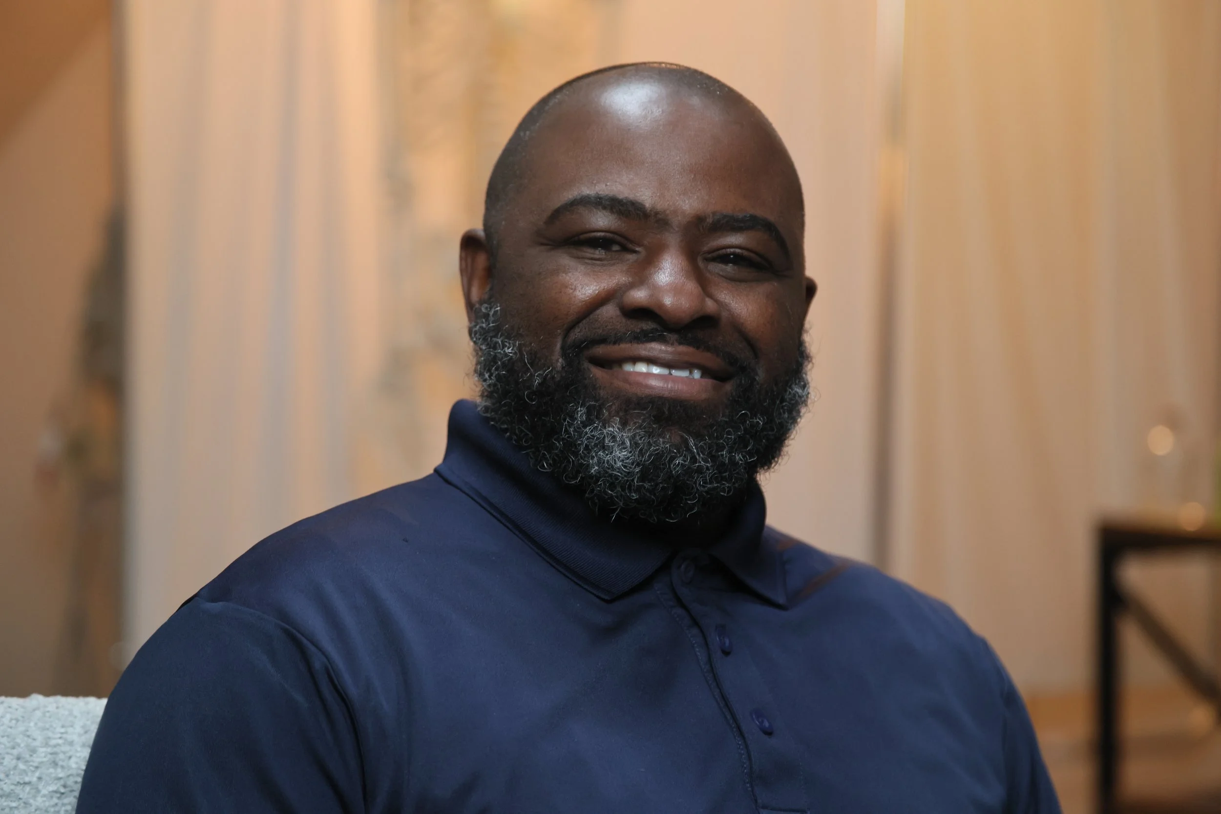 A smiling Black man with a beard wearing a dark blue polo shirt, sitting in a room with warm-colored curtains in the background.