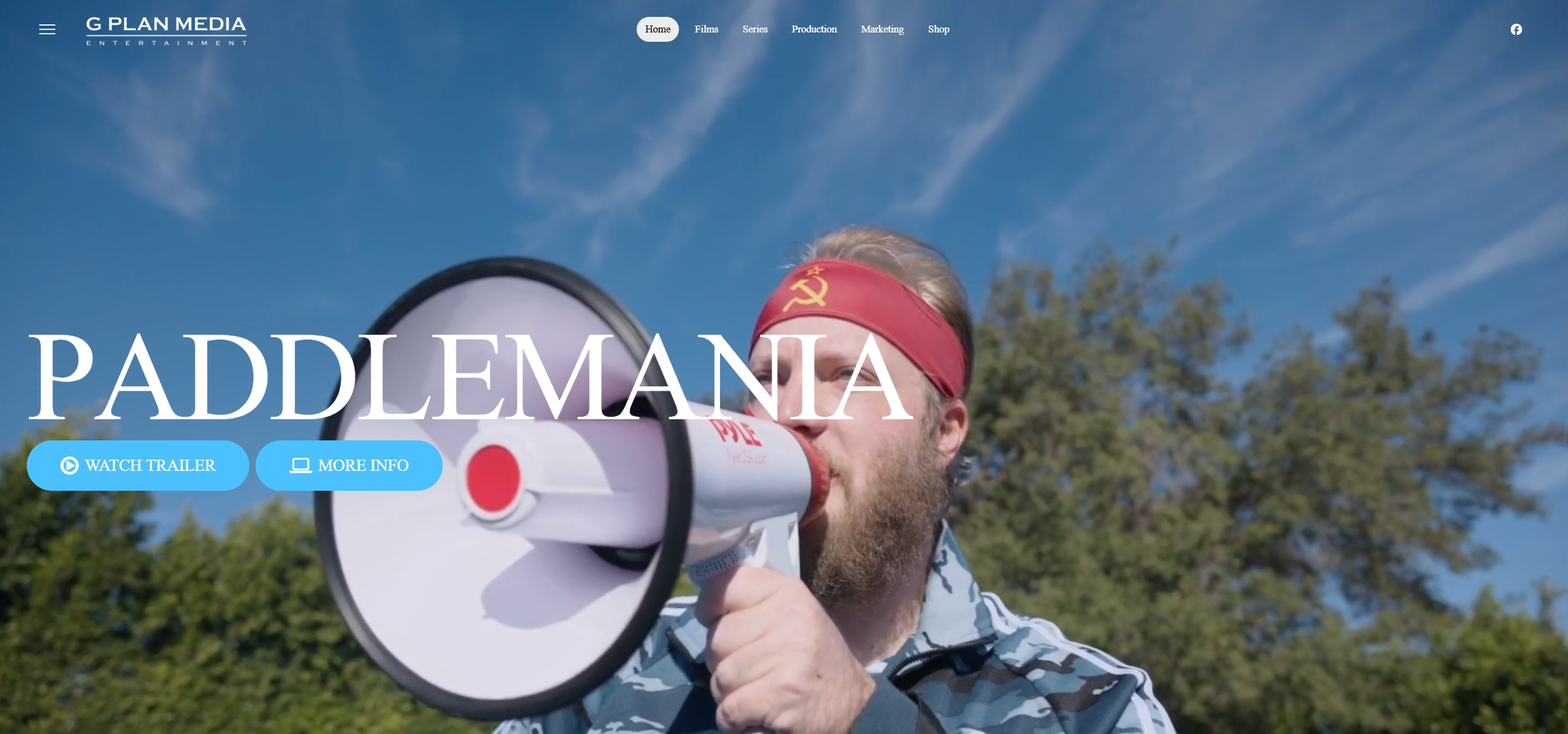 A man with a beard wearing a red headband with a hammer and sickle symbol, holding a megaphone to his mouth outdoors with trees and a blue sky in the background.