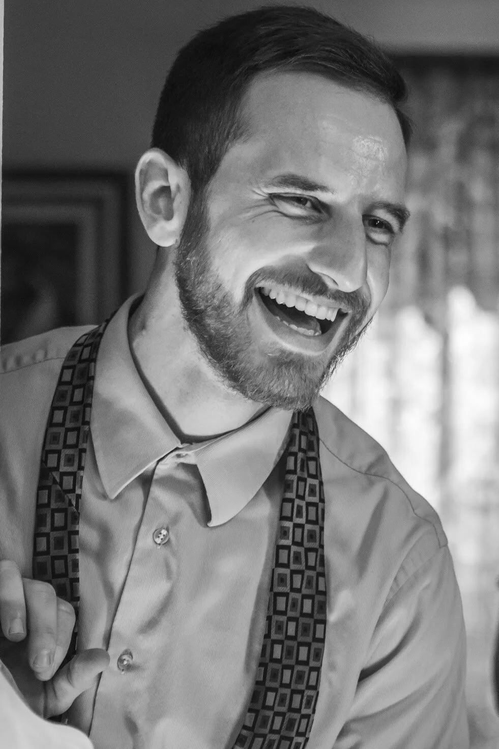Russel Donahue, smiling and laughing, wearing a button-up shirt and suspenders, indoors with blurred background.
