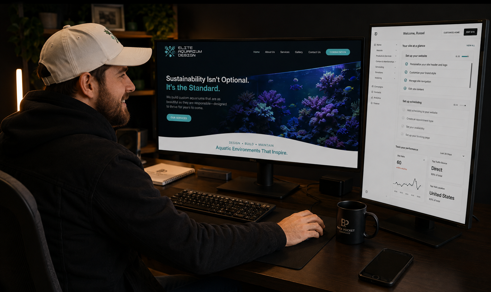 Russel Donahue with a white cap and dark hoodie sitting at a desk, working on dual monitors displaying a website and analytics dashboard. There is a black mug, a smartphone, a keyboard, and notebook on the desk.