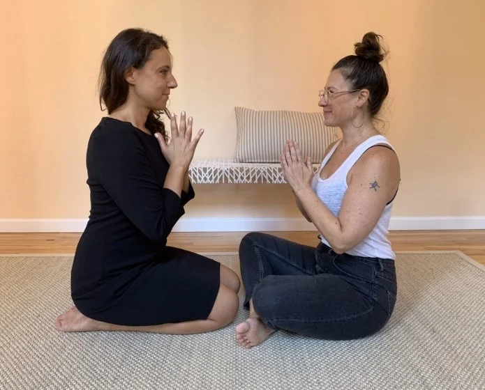 Two women sit on a beige rug facing each other with palms pressed together in a gesture of greeting or meditation, in a room with a wooden floor and a beige wall.