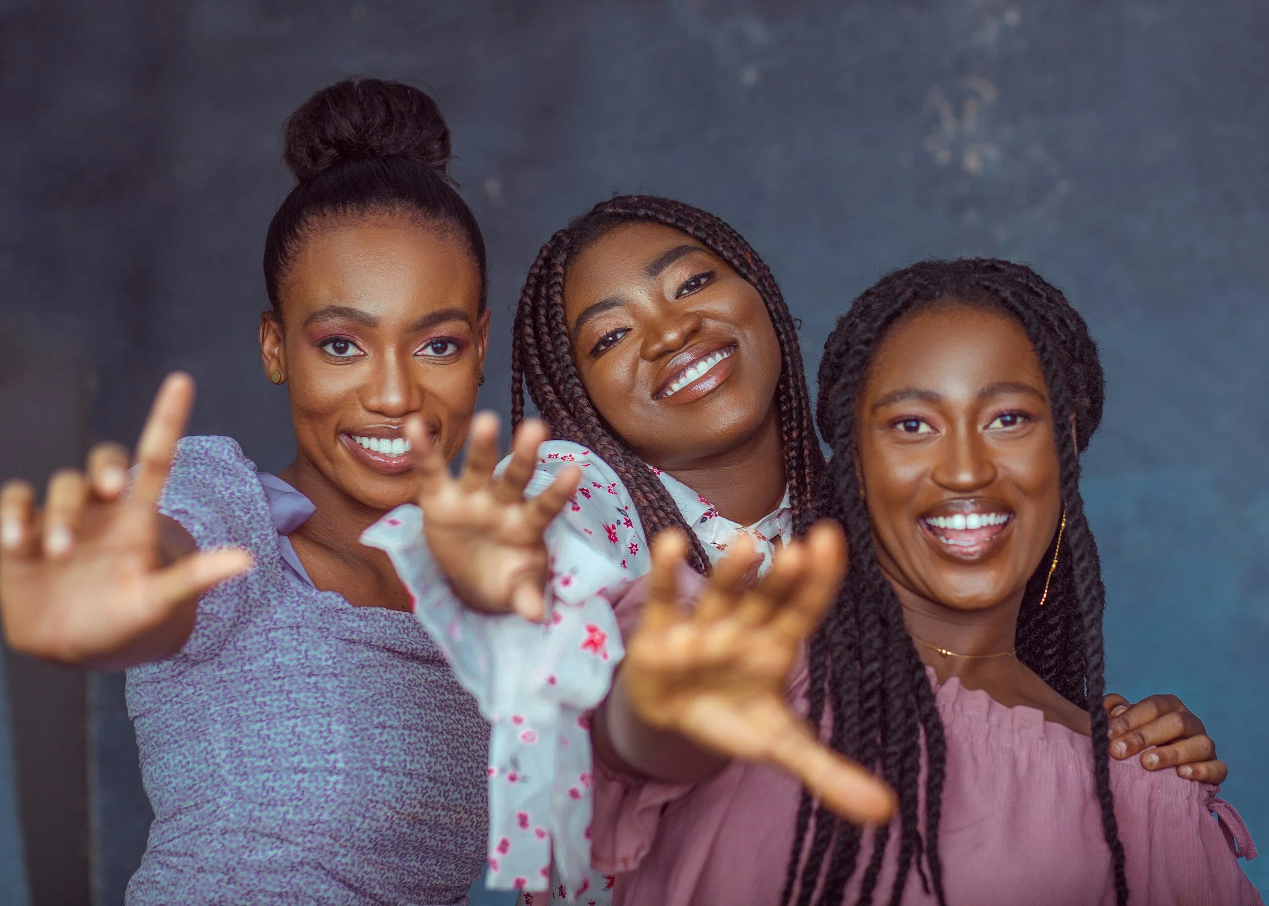 Three women smiling and reaching towards the camera, standing close together against a dark background.