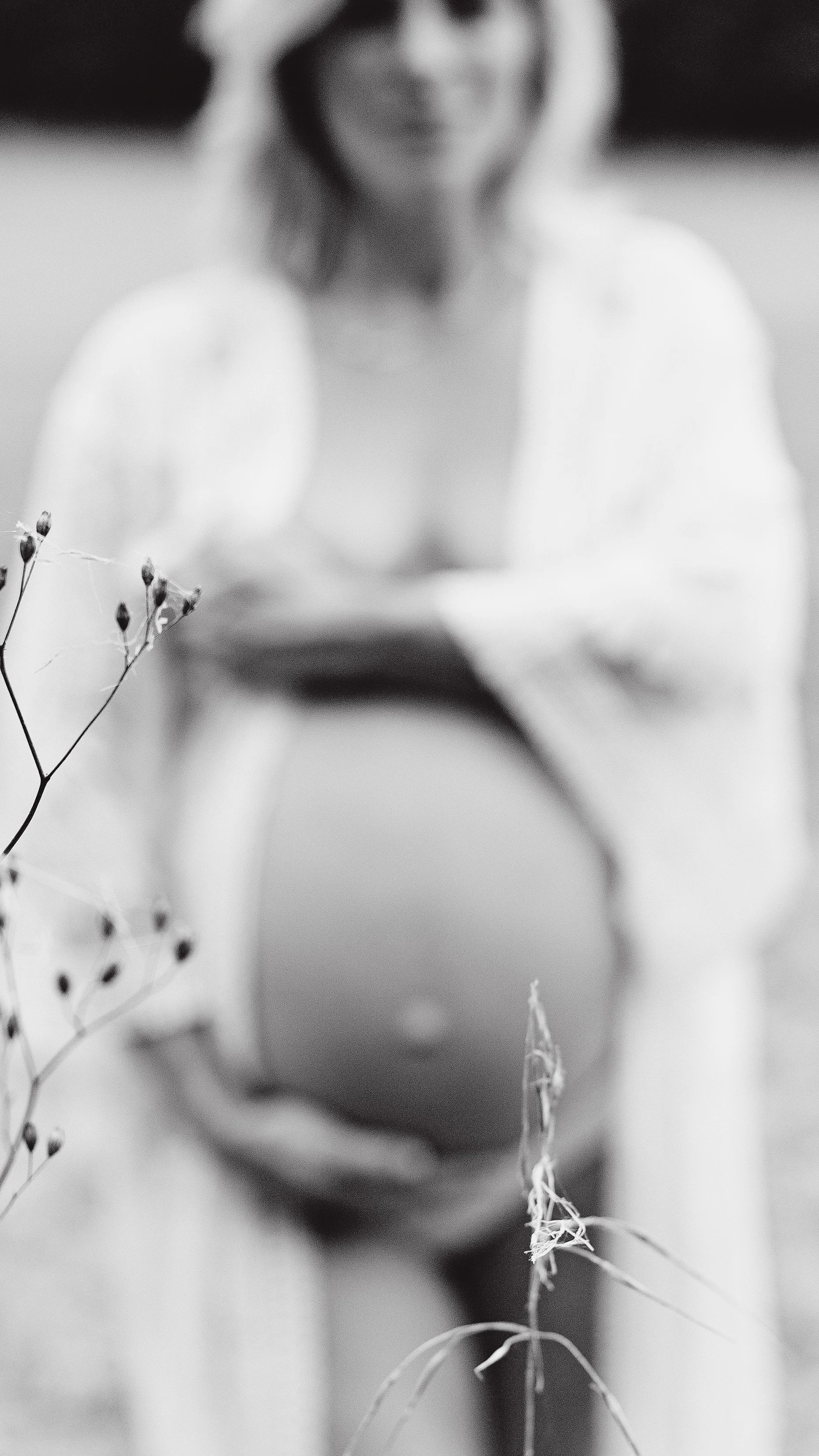 A pregnant mum cuddling her bump, the bump is visible and she is wearing a boho gown. The image is focused on a pretty flower with the mum out of focus.