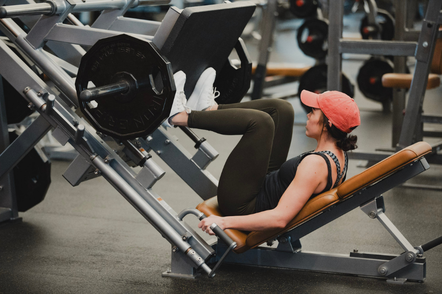 A woman in athletic wear and a red cap uses a leg press machine at a fitness center, focusing on strength training in a well-equipped gym.