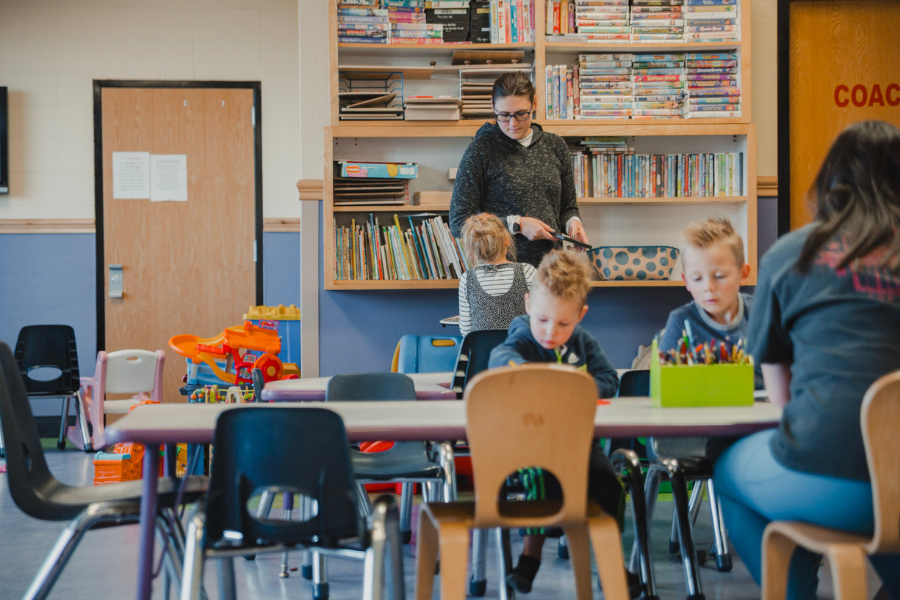 A group of young children engages in supervised activities at a child care facility, with books, toys, and art supplies in a bright, organized room.