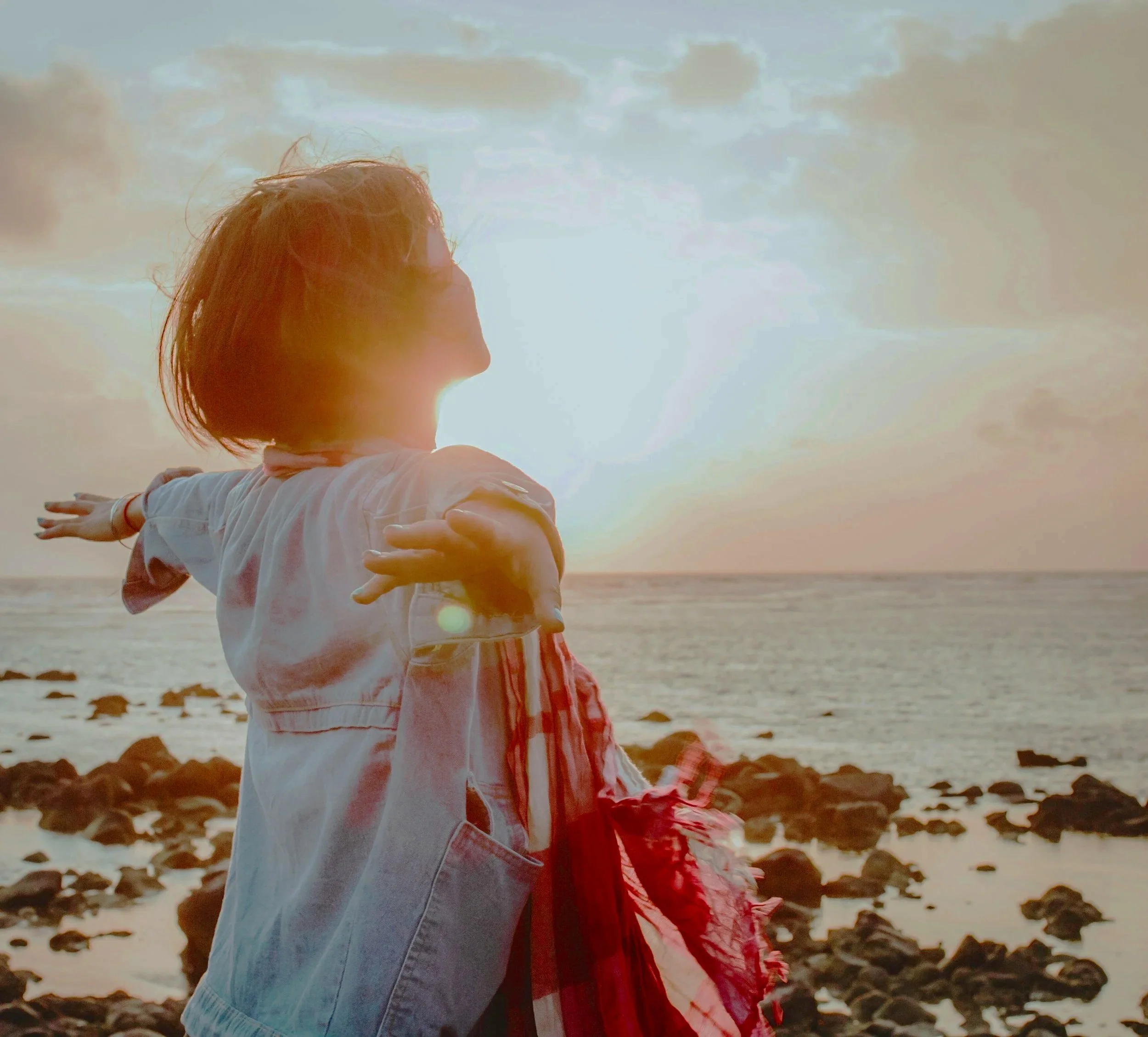 A person stands with ams out, looking at the sun over the ocean, feeling whole and healed.