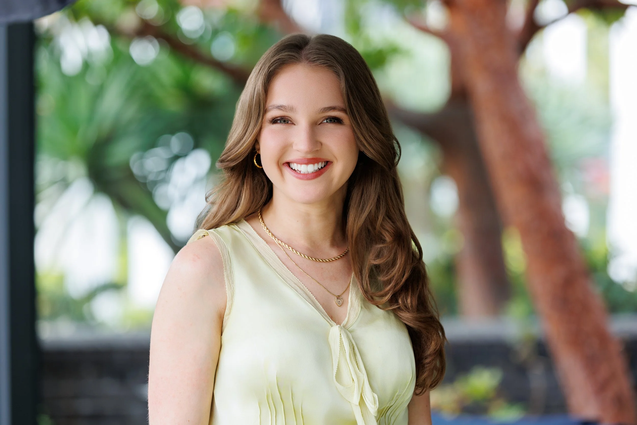 A woman with long brown hair smiling, wearing a sleeveless light yellow top, gold jewelry, and standing outdoors with blurred green trees in the background.