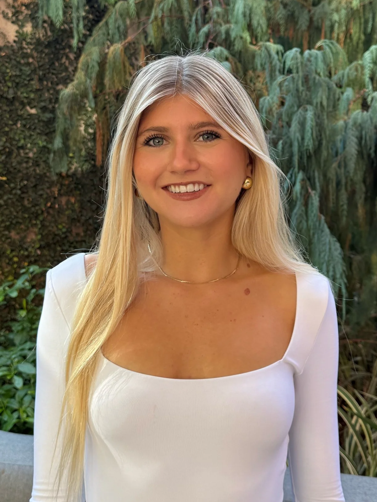 Smiling woman in business attire with long brown hair, standing outdoors with greenery in the background.