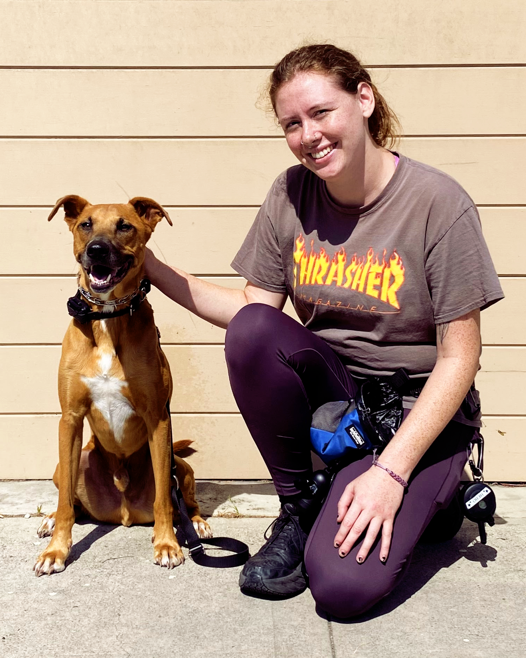 A young white woman with curly red hair smiling and kneeling next to a brown dog with a black collar, in front of a beige wooden wall.