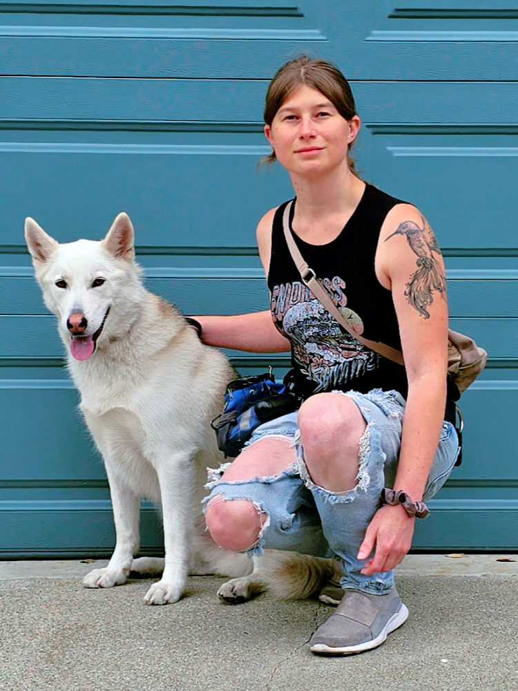 A young woman with tattoos on her arm, wearing a black tank top, ripped jeans, and gray sneakers, kneels beside a white dog with blue eyes and pointed ears, in front of a blue garage door.