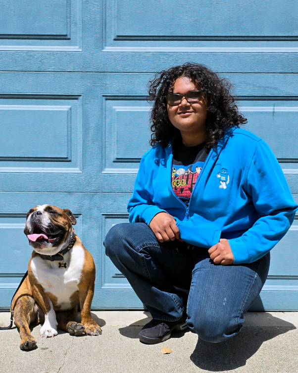 Hispanic woman with curly hair wearing sunglasses, a blue jacket, a graphic t-shirt, and jeans, crouching next to a happy, sitting bulldog with a collar, outside in front of a blue garage door.