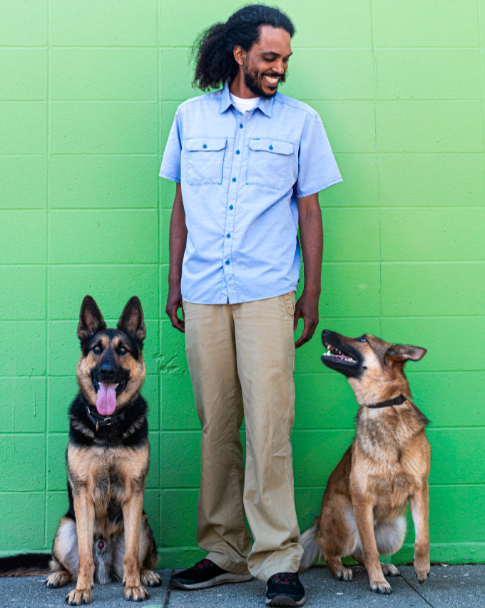 A man with dark curly hair wearing a light blue short-sleeve button-up shirt and khaki pants smiling at two dogs sitting next to him in front of a green wall.